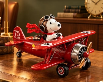 May include: A red biplane model featuring a Snoopy figurine wearing a brown aviator cap and goggles with a red scarf. The tail of the plane displays the text 75 YEARS in white. The aircraft has a silver engine, gold propeller, and black wheels.
