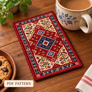 Puede incluir: Un posavasos rectangular con un patrón de punto de cruz que presenta un motivo de diamante azul central rodeado de diseños florales geométricos en rojo, crema y verde. El borde está compuesto por pequeños cuadrados azules y rojos. El texto PDF PATTERN es visible.