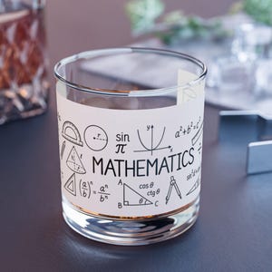 May include: A clear glass tumbler featuring a white wrap-around design printed with black mathematical symbols, geometric shapes, and the word MATHEMATICS in a bold, sans-serif font. The design includes a protractor, circle, and formulas.
