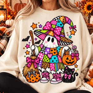 May include: A cream-coloured sweatshirt featuring a graphic of a ghost wearing a colourful patchwork witch hat and dress, holding a broom and a pumpkin bucket. The design includes a black cat, small stars, bats, and pink bows.