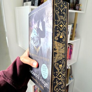 May include: A paperback book titled Clockwork Angel by Cassandra Clare, featuring a dark cover with a person in a suit and a gold-painted fore-edge with intricate filigree and butterfly designs, plus a blue circular sticker stating bonus content.