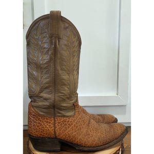 May include: A tall western-style boot featuring a textured, light brown foot with a distinct pebbled pattern and a smooth, medium brown shaft. The shaft is decorated with intricate, yellow-toned stitching in a leaf-like design and pull tabs.