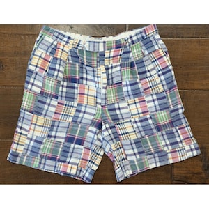 May include: A pair of shorts featuring a patchwork design made of various plaid fabric squares in shades of blue, green, yellow, pink, and white. The shorts include a waistband with a single white button closure and a front zipper fly.