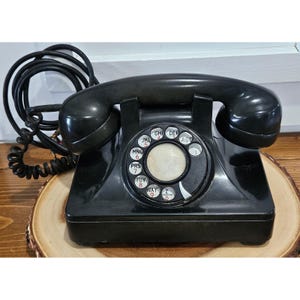 Puede incluir: Un teléfono de disco negro vintage con un auricular que descansa sobre una base con un dial circular. El dial incluye los números del cero al nueve, letras y la palabra OPERATOR. Un cable negro en espiral está unido al lado de la base.