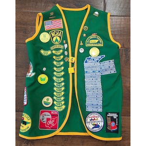 May include: A green vest with yellow trim, adorned with numerous embroidered patches and metal pins. The patches feature text including NCHA, Tennessee N.C.H.A. State Campouts, and various years from 1979 to 1991. An American flag patch is displayed on the upper left.