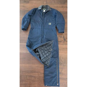 Könnte beinhalten: Ein dunkelblauer, isolierter Overall mit dunkelgrauem Kragen, Frontreißverschluss und Brusttaschen. Das Kleidungsstück hat ein schwarzes Steppfutter, das im geöffneten Beinbereich sichtbar ist, sowie einen durchgehenden seitlichen Reißverschluss.