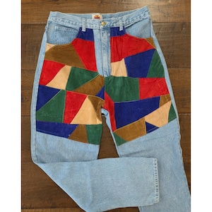 May include: Light blue denim jeans featuring a high-waisted design with a front panel composed of multi-colored suede geometric patches in red, blue, green, and tan. The jeans include a metal button closure and a small brand label at the waist.