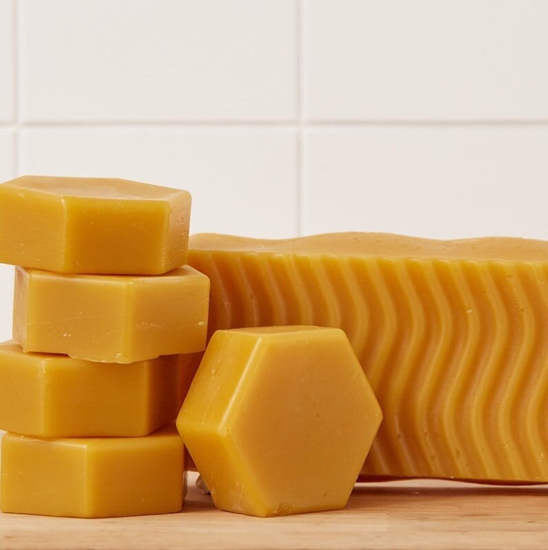 May include: A stack of hexagonal beeswax blocks and a larger rectangular block of beeswax. The beeswax is a pale yellow color.