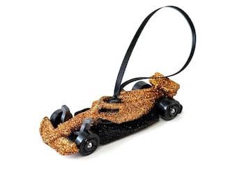 May include: A glittery gold and black race car ornament featuring a sleek aerodynamic body, four black wheels, and a black ribbon loop attached to the top for hanging. The car has a textured, sparkling finish on the chassis and spoiler.
