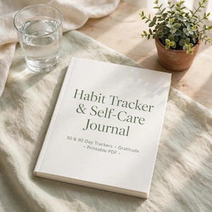 Pu&ograve; includere: Un diario bianco con il testo Habit Tracker & Self-Care Journal, 30 & 90 Day Trackers, Gratitude e Printable PDF stampato in verde scuro sulla copertina. Il diario &egrave; posizionato su una superficie in tessuto di colore chiaro.