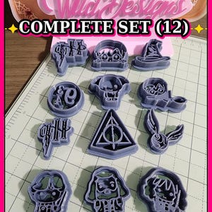 May include: A complete set of 12 purple plastic cookie cutters arranged in a grid on a cutting mat. The cutters feature various shapes including a lightning bolt, a wizard hat, a triangle symbol, and character outlines. A sign reads Wild Designs COMPLETE SET (12).