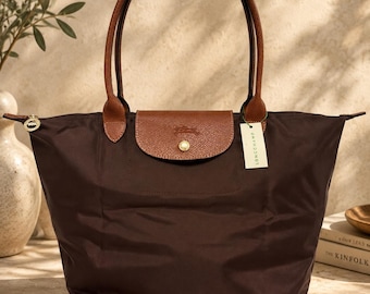 May include: A dark brown nylon tote bag featuring two brown leather handles and a brown leather flap closure with a gold-tone snap button. A white tag with the text LONGCHAMP is attached to the handle. The bag has a zipper closure.