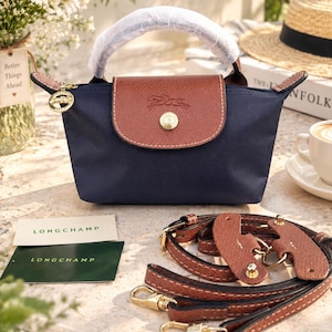 May include: A navy blue handbag with a brown leather flap featuring a gold-tone snap button and a horse logo. The bag has a top handle wrapped in protective paper, a detachable brown leather shoulder strap, and two Longchamp brand cards.