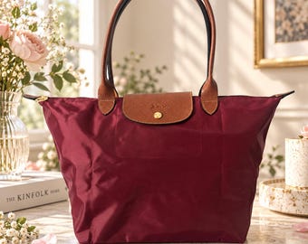 May include: A deep burgundy nylon tote bag featuring brown leather handles and a matching brown leather flap closure with a gold-toned snap button. The bag has a structured silhouette with a wide base and narrow top, showcasing a smooth, durable fabric finish.