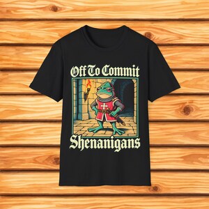 May include: A black t-shirt featuring a graphic of a green frog dressed as a medieval knight in a red tunic with a white cross, holding a sword and a torch. The text above reads Off To Commit and the text below reads SHENANIGANS in a bold font.