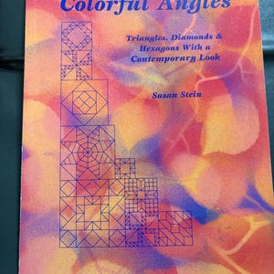 Puede incluir: Un libro titulado Colorful Angles de Susan Stein con el subtítulo Triangles, Diamonds & Hexagons With a Contemporary Look. La portada presenta un fondo de acuarela vibrante en naranja, rojo y morado con patrones geométricos de líneas azules.