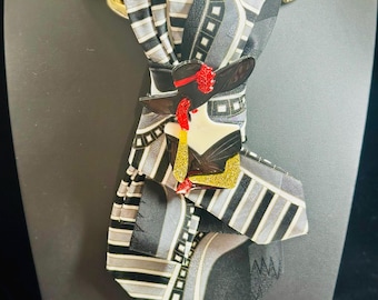 Puede incluir: Una corbata con un patrón geométrico en negro, blanco y gris, acentuada por un collar de cadena dorada. Un broche decorativo con forma de figura que lleva un sombrero negro de ala ancha con detalles en rojo y purpurina dorada está prendido en el centro.