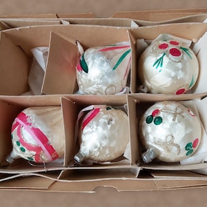 May include: A set of five glass Christmas ornaments nestled in a cardboard box with dividers. The ornaments are cream-colored with a pearlescent finish, featuring hand-painted red and green floral motifs, gold glitter accents, and silver metal caps.