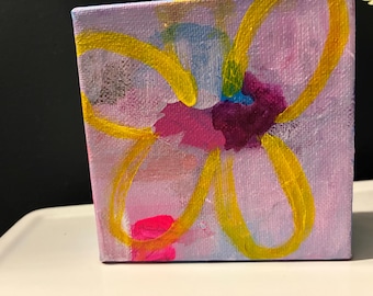 May include: A square canvas painting featuring a light purple background with a stylized flower outline in bright yellow paint. The center of the flower contains abstract patches of dark purple, magenta, and light blue acrylic paint strokes.