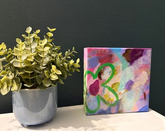 May include: A square canvas painting featuring an abstract background of blended pink, purple, blue, and yellow paint strokes with a prominent, hand-painted bright green flower outline centered on the left side of the canvas surface.
