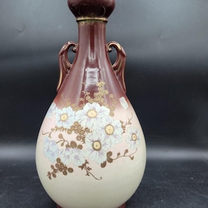 May include: A ceramic vase with a bulbous body and a narrow neck featuring a dark maroon top and base that fades into a cream-colored center. The vase is decorated with white floral patterns, gold-colored accents, and two small side handles.