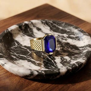 May include: A gold-toned ring featuring a large, rectangular, faceted deep blue gemstone. The band of the ring displays a textured, cross-hatched pattern on the sides, providing a distinct contrast to the smooth, polished metal setting.