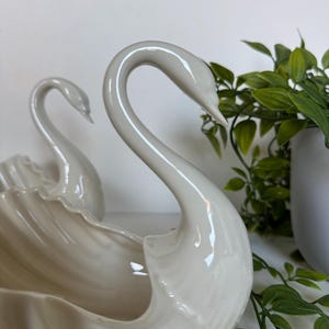 May include: Two glossy off-white ceramic swan-shaped bowls featuring curved necks and molded wing details. The swan figures have smooth, reflective surfaces and are positioned one behind the other, creating a decorative set of containers.