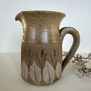 May include: A ceramic pitcher with a speckled tan upper section and a darker brown lower body. The lower portion features a repeating pattern of white leaf imprints and thin dark stalks. It has a curved handle and a small pouring spout.