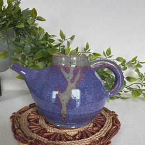 May include: A ceramic teapot with a glossy finish, featuring a deep purple body with a speckled texture and a prominent gray vertical drip pattern extending from the rim. It has a curved handle and a tapered spout, resting on a woven mat.