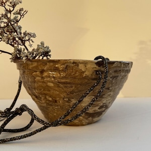 May include: A round, hand-formed ceramic hanging planter with a marbled brown and beige glaze. The bowl features a textured, uneven surface and is suspended by dark, braided nylon cords. A small, dried plant sprig rests inside the rim.