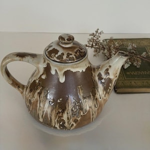 May include: A ceramic teapot featuring a brown base with a white drip glaze pattern. The teapot has a matching lid with a small knob handle, a curved spout, and a side handle. The glaze creates a mottled effect over the brown surface.