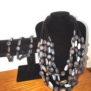 May include: A multi-strand necklace and a matching bracelet featuring a combination of dark gray textured porous beads and elongated, iridescent blue-gray shell-like beads strung together with small black spacer beads.