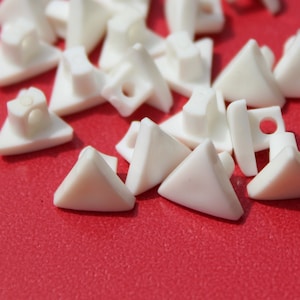 May include: A group of white plastic triangular buttons with a hole in the center. The buttons are scattered on a red surface.