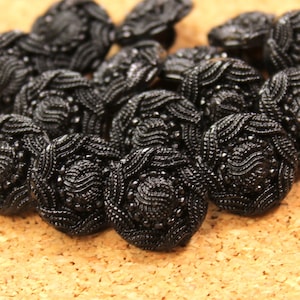 May include: A group of black decorative buttons with a floral pattern. The buttons are clustered together, displaying their detailed woven texture and small, shiny embellishments. They are placed on a light brown, textured surface.