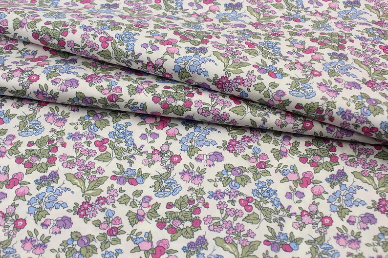 May include: A white fabric featuring a dense, repeating floral pattern with small pink, purple, and light blue flowers interspersed with green leaves and stems. The textile has a soft, folded texture, showcasing the intricate botanical print throughout.
