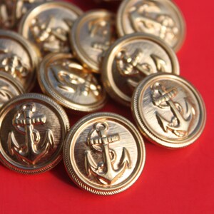 May include: A group of gold-colored metal buttons with an anchor design. The buttons are arranged in a pile on a red surface.
