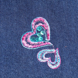 May include: Close-up of a dark blue denim fabric with an embroidered heart design. The design features two connected hearts, one larger than the other, with pink and teal stitching and teal sequins.