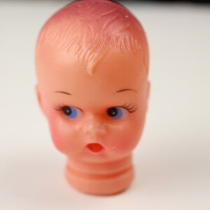 May include: A vintage doll head with a peach complexion, blue eyes, and delicate eyelashes. The doll head has a surprised expression with rosy cheeks and a small, open mouth. The hair is styled with a slight wave.
