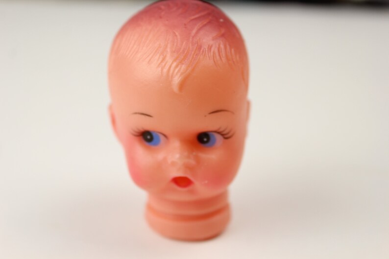 May include: A vintage doll head with a peach complexion, blue eyes, and delicate eyelashes. The doll head has a surprised expression with rosy cheeks and a small, open mouth. The hair is styled with a slight wave.