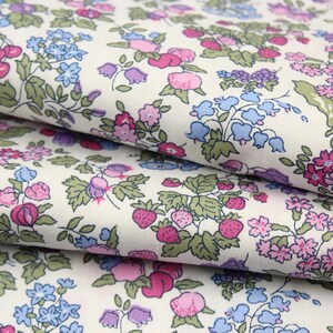 May include: A white fabric featuring a dense, repeating pattern of small pink strawberries, blue bell-shaped flowers, and clusters of pink and purple blossoms with green leaves. The material is folded in layers, showcasing the continuous floral print.