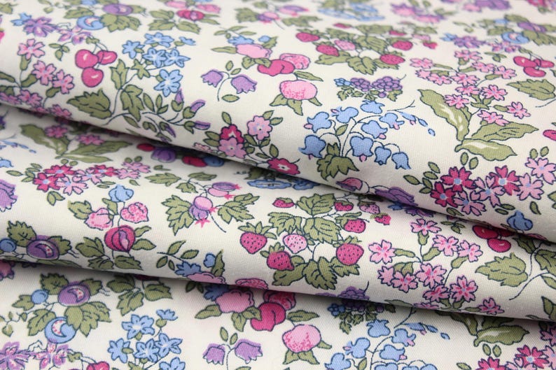 May include: A white fabric featuring a dense, repeating pattern of small pink strawberries, blue bell-shaped flowers, and clusters of pink and purple blossoms with green leaves. The material is folded in layers, showcasing the continuous floral print.
