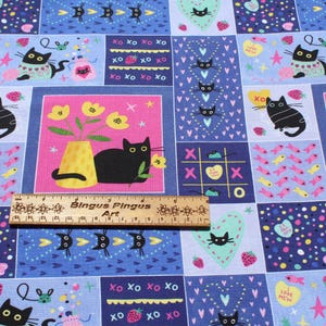 May include: A colorful fabric with a repeating pattern of black cats, flowers, hearts, strawberries, and the text "LOVE MEW". The design includes pink, blue, and yellow squares with various cat-themed illustrations. A wooden ruler is placed on the fabric.