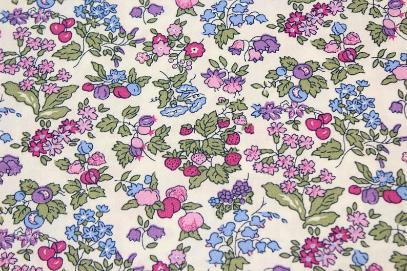 May include: A cream-colored fabric featuring a dense, repeating botanical print of small pink, purple, and blue flowers, green leaves, and clusters of red strawberries and cherries scattered across the surface in a detailed, colorful pattern.