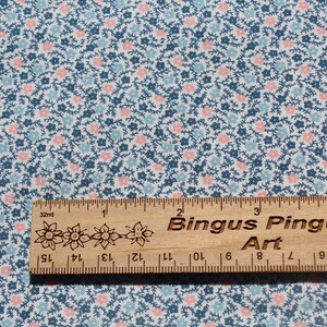 May include: Fabric with a small floral pattern in blue, pink, and white. A wooden ruler with the text "Bingus Pingus Art" is placed on the fabric. The ruler is marked in inches, with measurements in centimetres.
