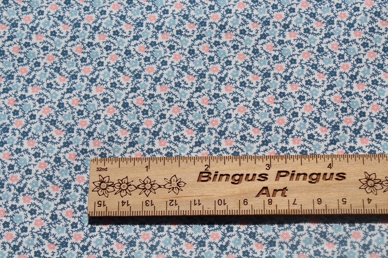 May include: Fabric with a small floral pattern in blue, pink, and white. A wooden ruler with the text "Bingus Pingus Art" is placed on the fabric. The ruler is marked in inches, with measurements in centimetres.