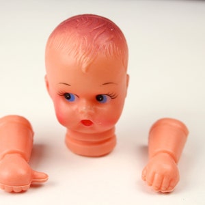 May include: A disassembled vintage doll head and arms. The doll head is peach-colored with blue eyes, red cheeks, and a surprised expression. The arms are also peach-colored, with one hand open and the other clenched.