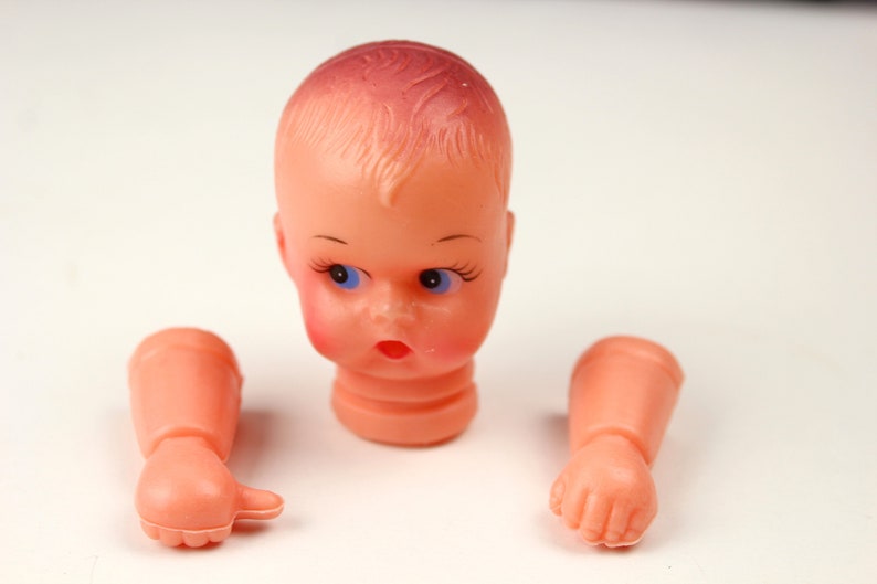 May include: A disassembled vintage doll head and arms. The doll head is peach-colored with blue eyes, red cheeks, and a surprised expression. The arms are also peach-colored, with one hand open and the other clenched.