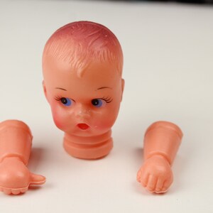 May include: A disassembled vintage doll head and arms. The doll head is peach-colored with blue eyes, rosy cheeks, and a red mouth. The arms are also peach-colored, with one hand open and the other in a fist. The doll parts are on a white background.