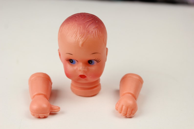 May include: A disassembled vintage doll head and arms. The doll head is peach-colored with blue eyes, rosy cheeks, and a red mouth. The arms are also peach-colored, with one hand open and the other in a fist. The doll parts are on a white background.