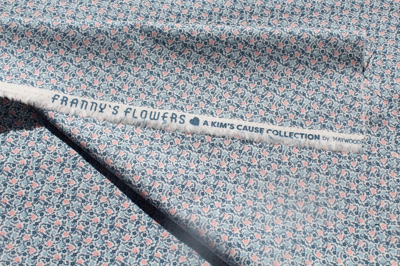 May include: Fabric with a floral pattern in blue, pink, and white. The fabric features a repeating design of small flowers and hearts. A white strip with the text "FRANNY'S FLOWERS A KIM'S CAUSE COLLECTION by MAYWOOD" is visible.